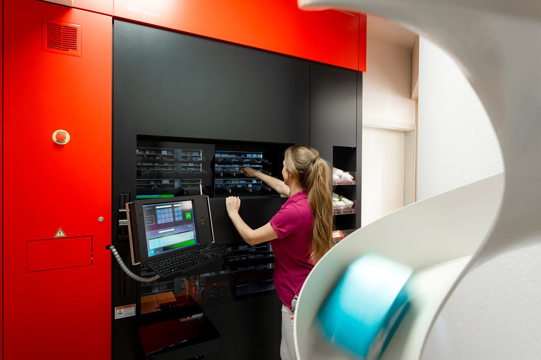 Fotografie einer Frau, die an einem großen, roten und schwarzen automatisierten Lager- oder Verpackungssystem arbeitet, mit mehreren Fächern und einem Computerbildschirm zur Steuerung. 