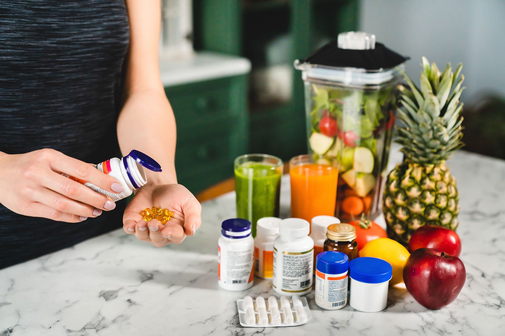 Fotografie zeigt Person, die Nahrungsergänzungsmittel in Kapselform aus einer Flasche in Hand nimmt, vor mehreren Vitamin- und Supplementbehältern sowie frischem Obst und Gemüse auf Marmorarbeitsplatte.