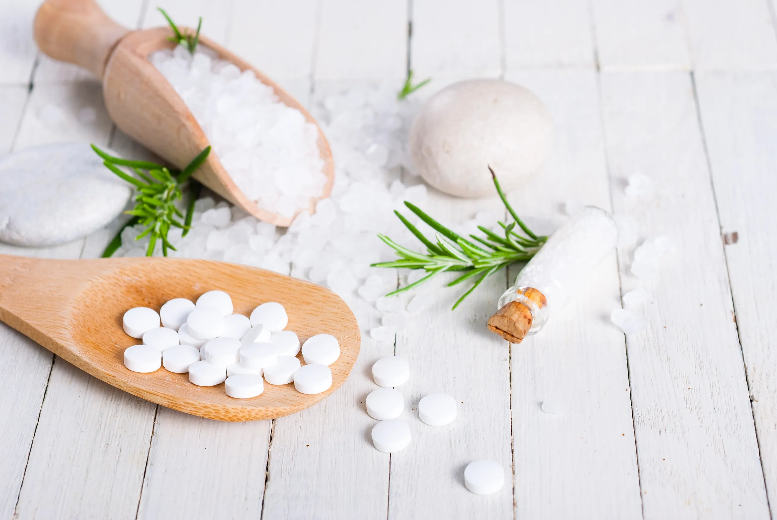 Fotografie zeigt weiße Tabletten auf Holzlöffel und verstreut auf weißem Holztisch, umgeben von grobem Salz, grünen Rosmarinzweigen und einem kleinen Glasfläschchen mit Korken.
